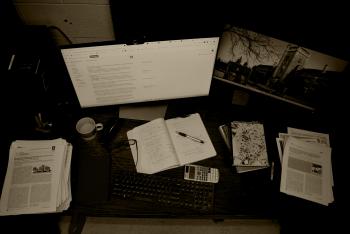 A messy table with a monitor, keyboard, mouse, copy, cup, pens, glasses, papers etc.
