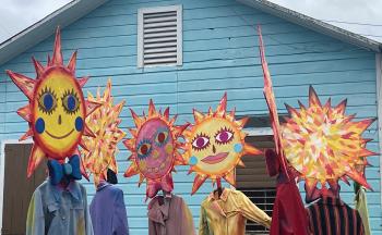 The photo shows seven colourful, handmade, sun-shaped puppets in front of a blue house.