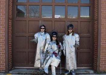 Three women standing in front of a garage wearing aluminum-rayon coats, spats, and kevlar gloves that give a sci-fi aesthetic 