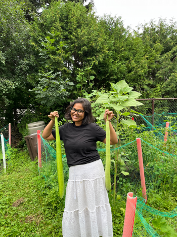A smiling woman holds two long, pale-green vegetables in a community garden surrounded by leafy plants and green mesh fencing.
