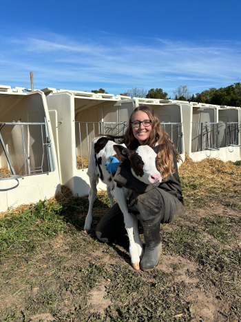 PhD student Lydia Conrad embracing a week-old Holstein heifer calf in front of a row of calf hutches, on a sunny day. 