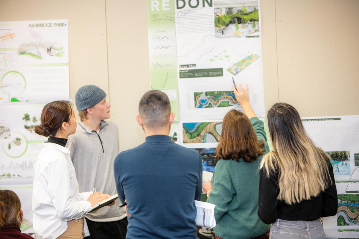   People gathered looking at project panels.  Professor pointing at the panel on the wall.