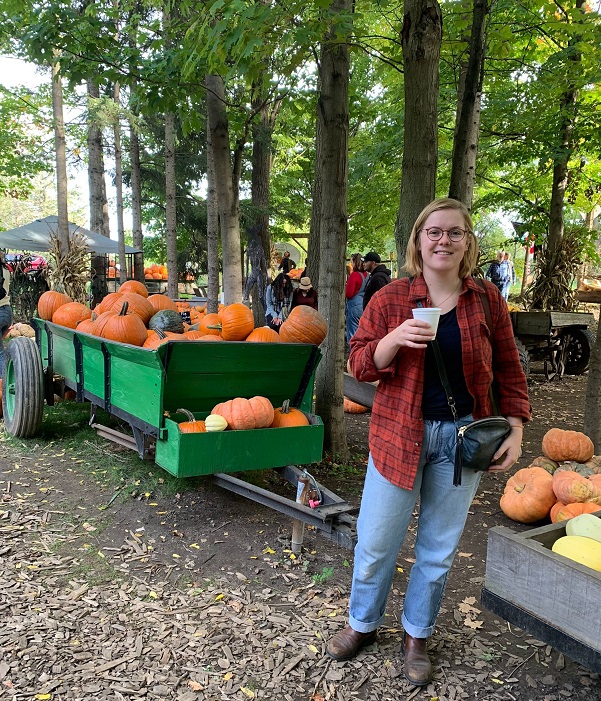 Regan Zink holding a drink in front of a wagon of pumpkins with trees in the background
