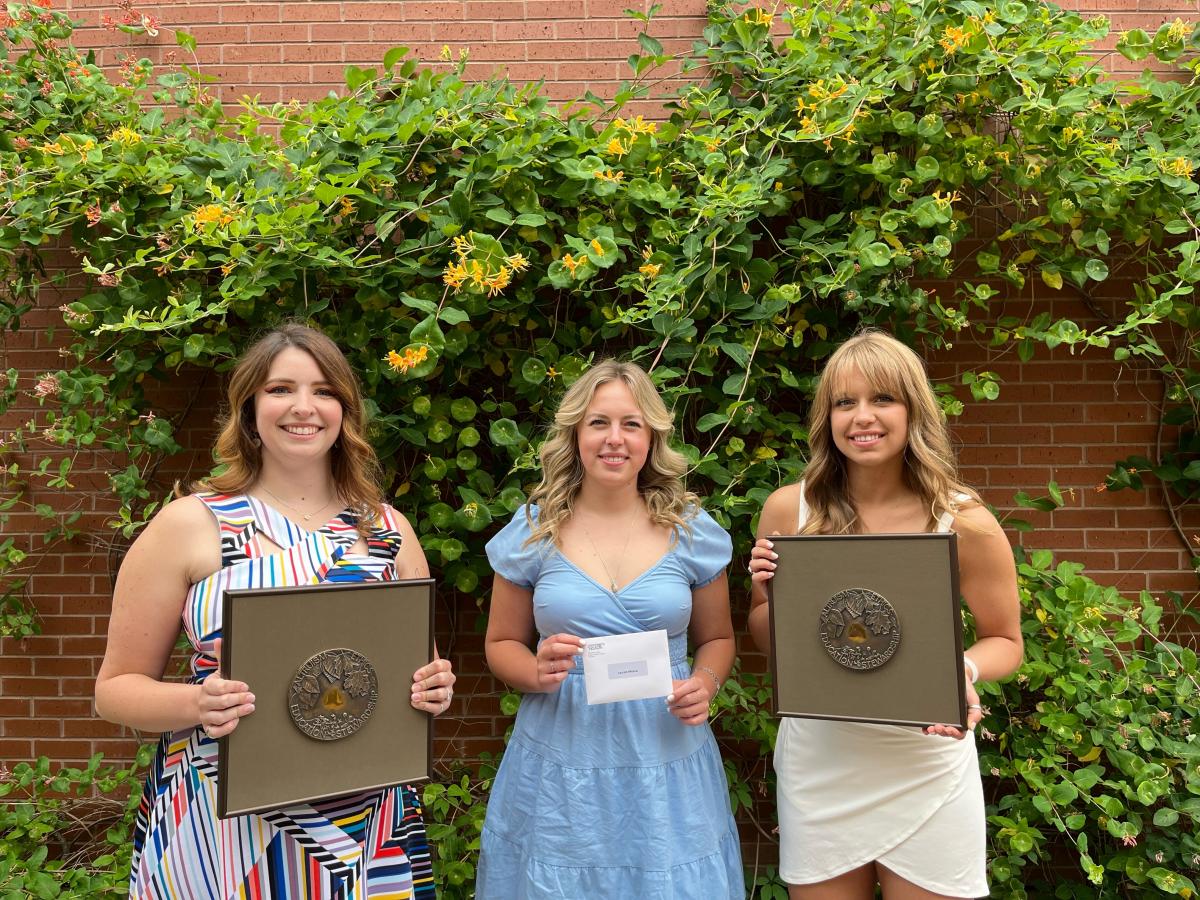 Three graduating students posing with their convocation awards against vine wall