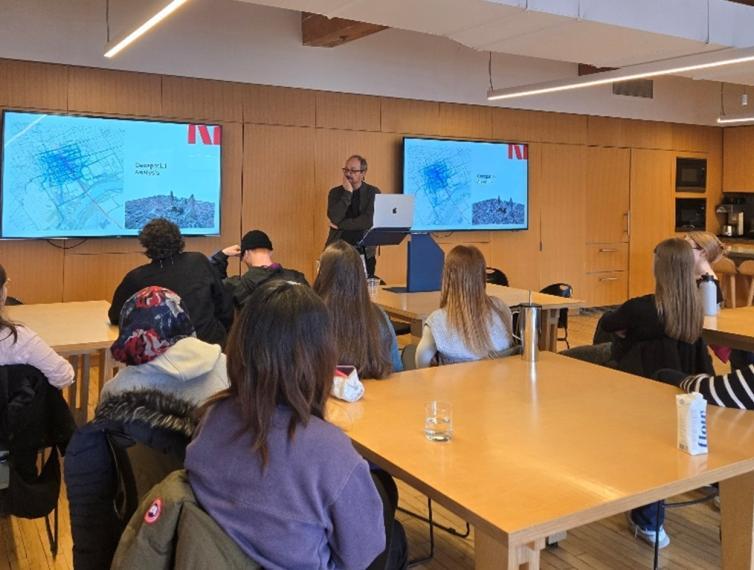students sitting at tables listening to a speaker at the front of the room