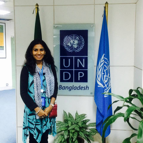 Faria in blue dress, scarf and black sweater stands in front of a UNDP sign