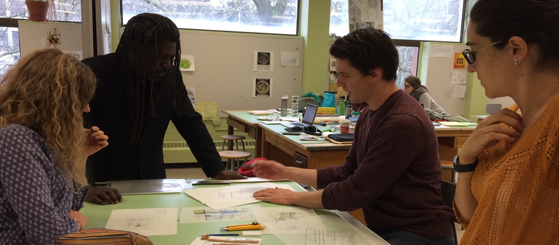 Kona Gray giving MLA desk crits to three students around drafting table