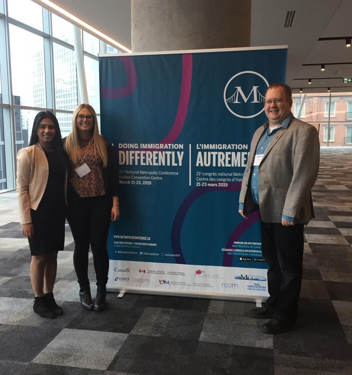 Three people in front of 2019 Metropolis Immigration Conference Banner